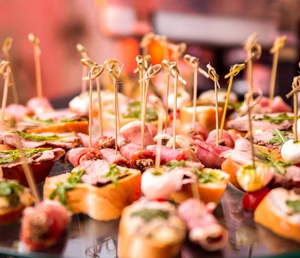 Assorted small appetizers on skewers, including meats, cheeses, and vegetables, garnished with herbs on a black platter.
