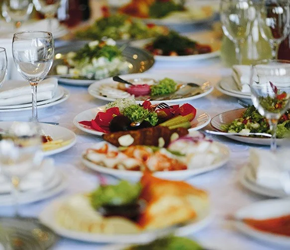 A table set with various plates of food including salads, meats, and side dishes, wine glasses, and utensils for a meal.