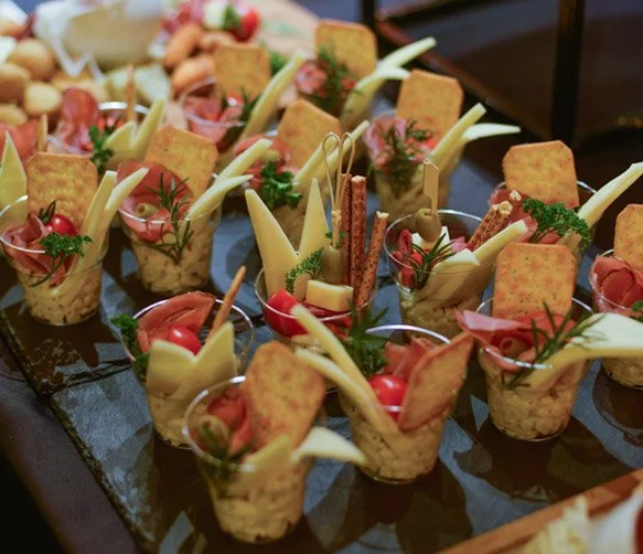 Assorted savory appetizer cups with cheese, cherry tomatoes, crackers, herbs, and slices of cured meat, arranged on a tray.