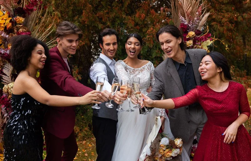 Six people at a wedding toast with champagne outdoors, smiling and celebrating.