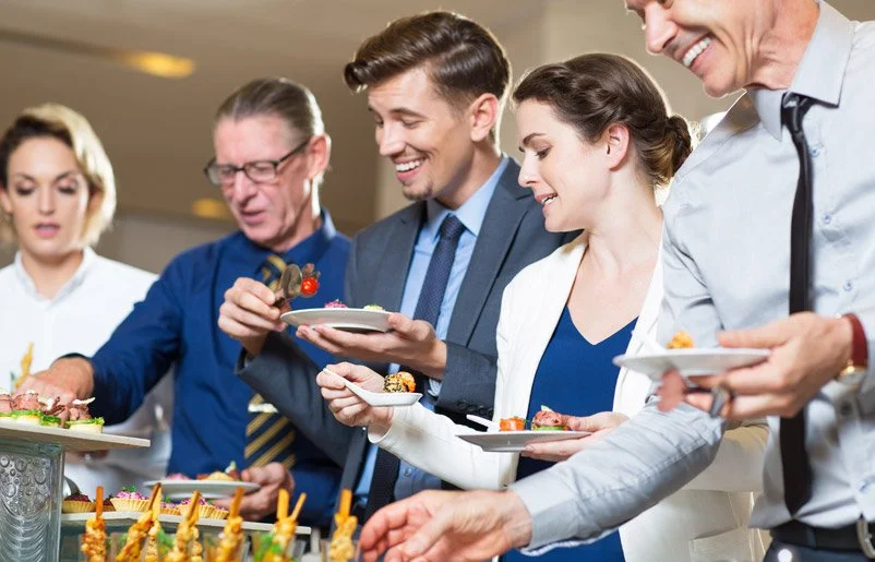 People in business attire at a buffet table serving themselves food and smiling.