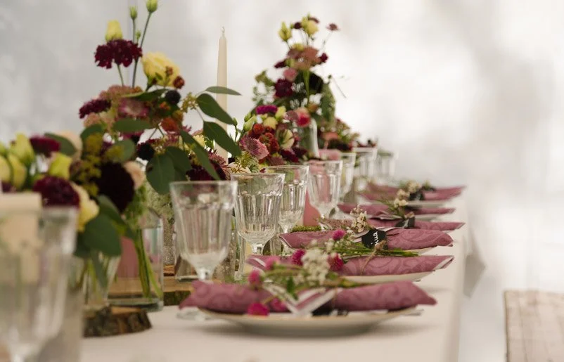 A decorated dining table with glassware, pink napkins, floral arrangements, and candles for a celebration or event.