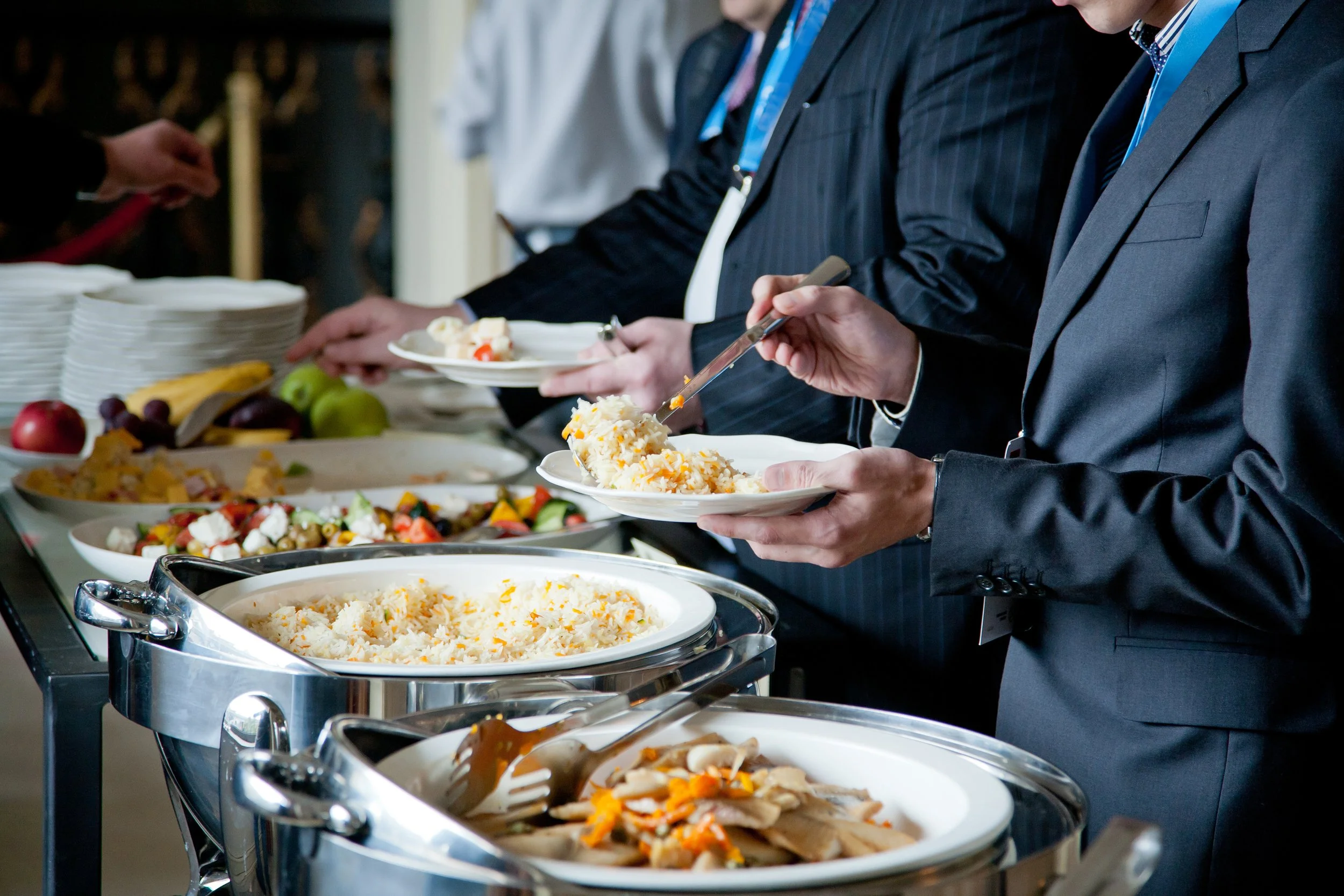 People dressed in formal business suits serving themselves from a buffet of rice, vegetables, and other dishes at a catered event.