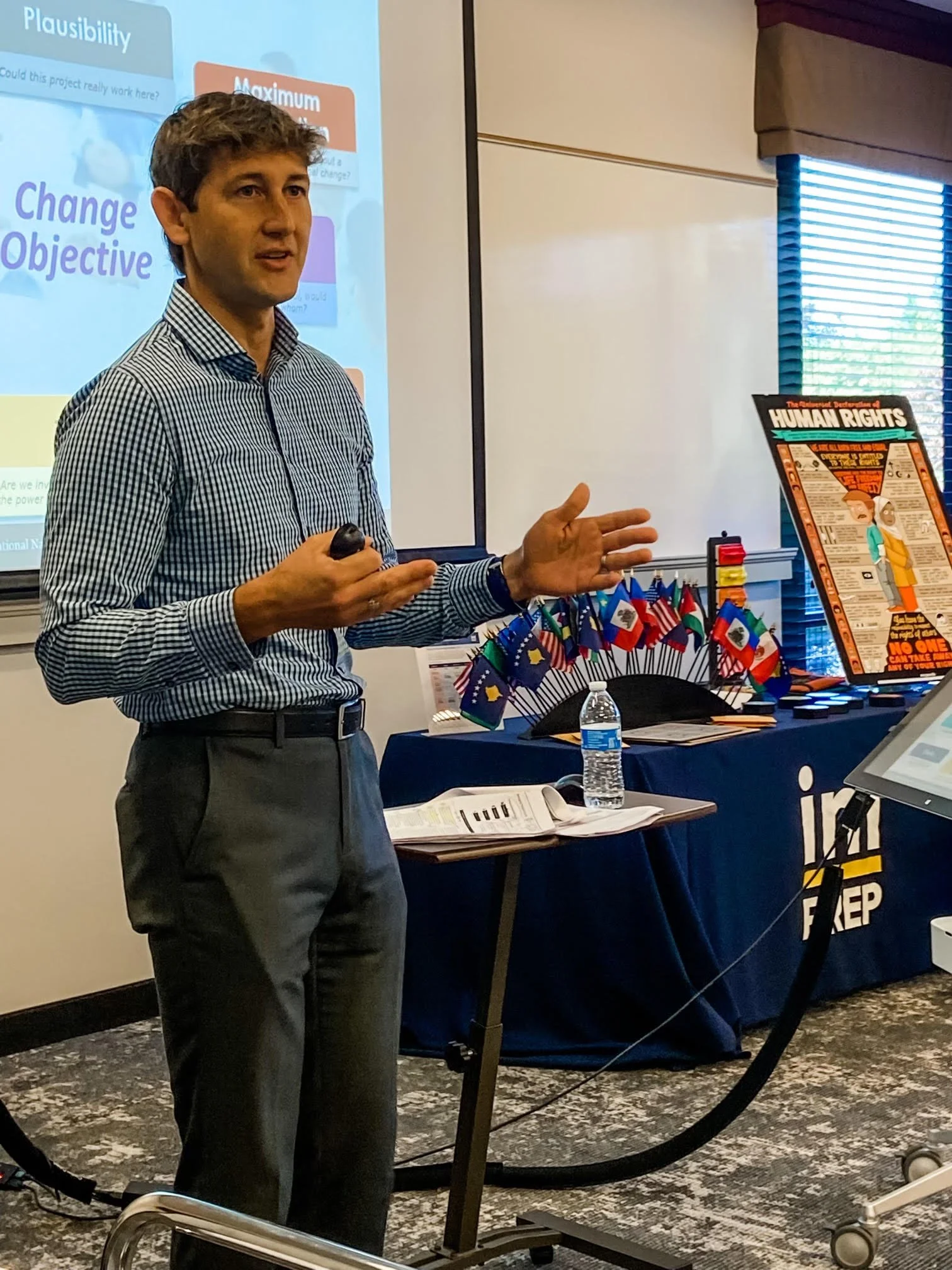 A man giving a presentation in a classroom, pointing with his left hand while holding a remote in his right, standing in front of a projection screen and a table with flags, books, and a water bottle.