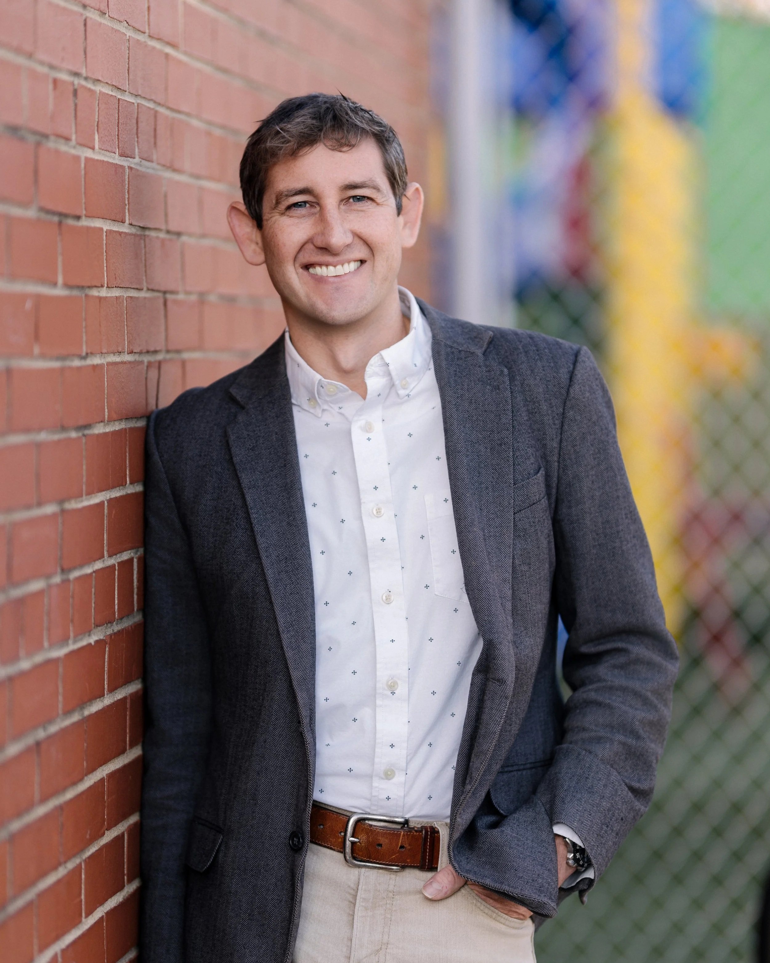 A man with brown hair and a smile, wearing a dark blazer and a white shirt, standing next to a brick wall outdoors.