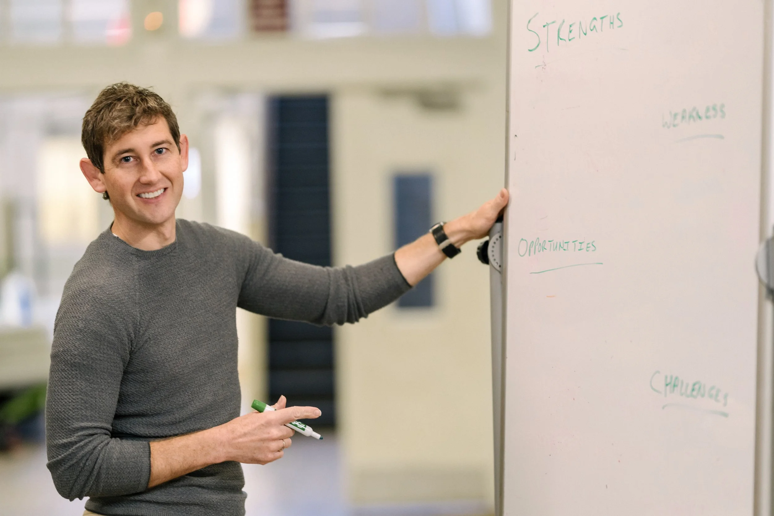 A man with short brown hair, wearing a gray sweater, standing next to a whiteboard with handwritten words, smiling and holding a green marker.