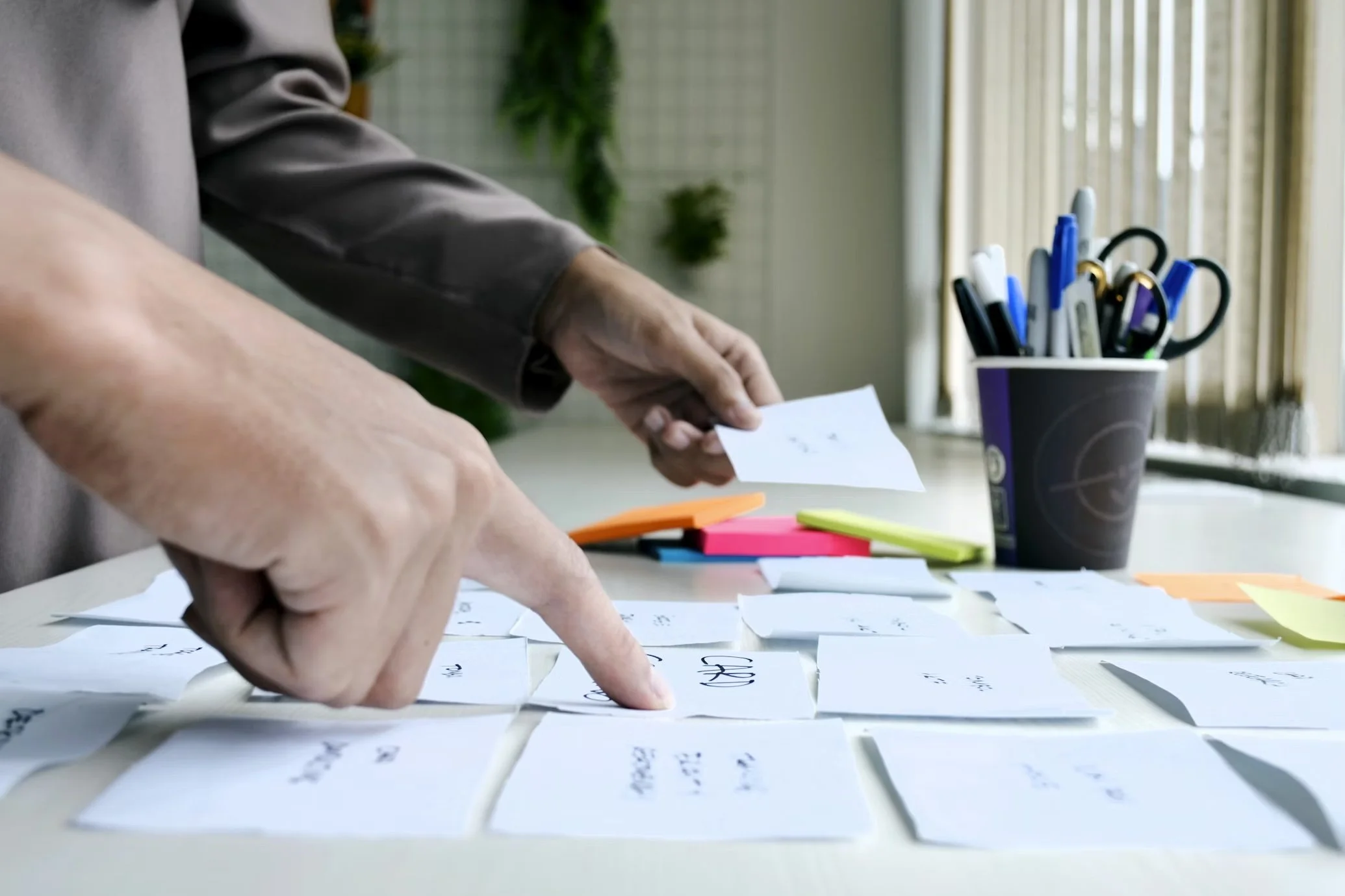 Person organizing notes and sticky notes on a table with pens and scissors in a paper cup nearby.