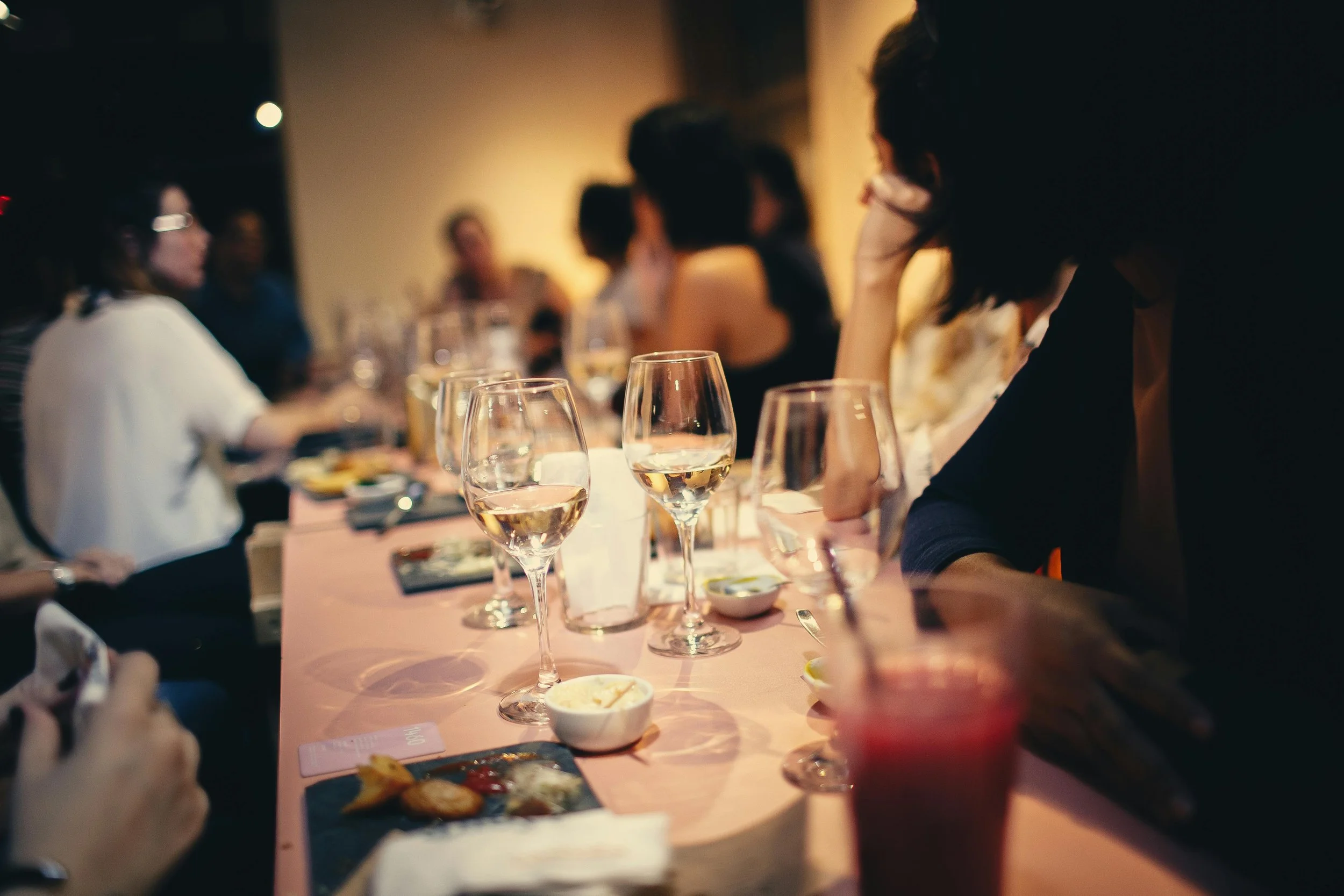 People sitting at a long dinner table during a social gathering, with glasses of white wine and various dishes on the table.
