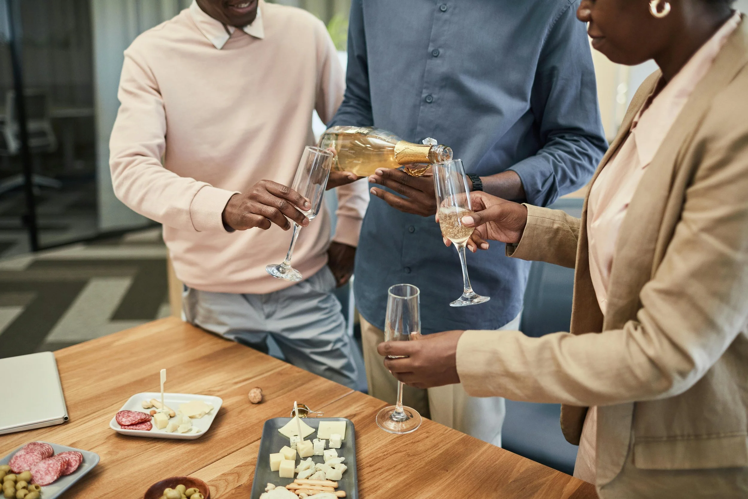 People pouring champagne into glasses during a gathering, with cheese and meats on the table.