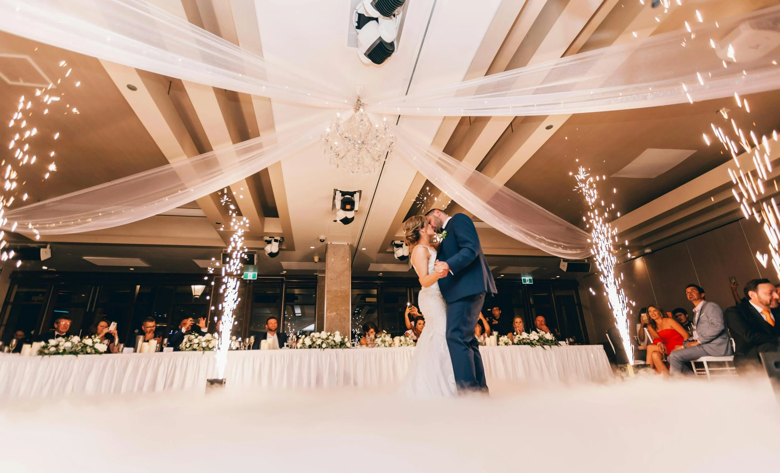A newlywed couple dancing at their wedding reception with guests seated at a long head table in the background. The venue is decorated with drapes, a chandelier, and sparklers creating a festive atmosphere.