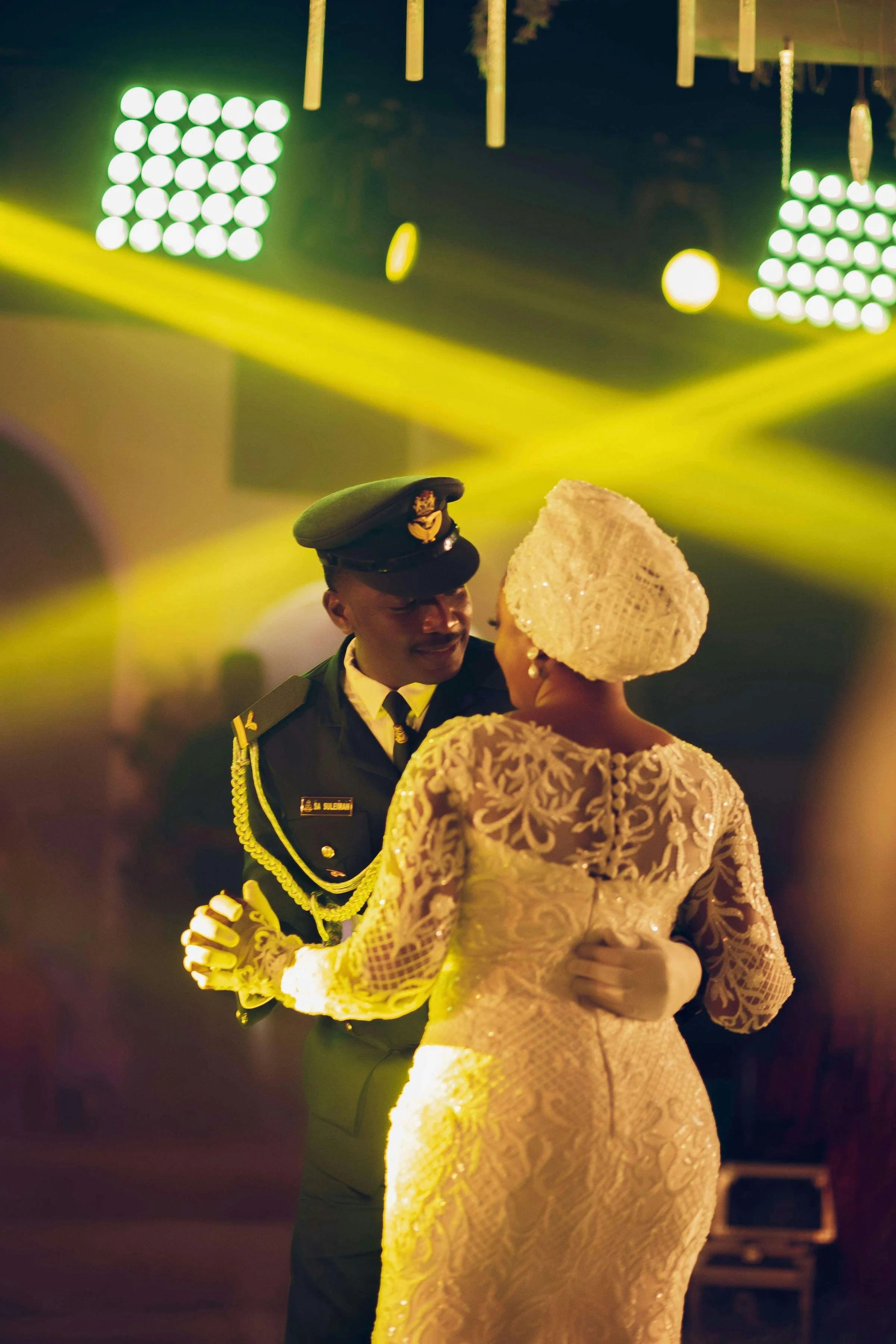 A couple dancing together, dressed in military and wedding attire, with yellow lights and lasers in the background.