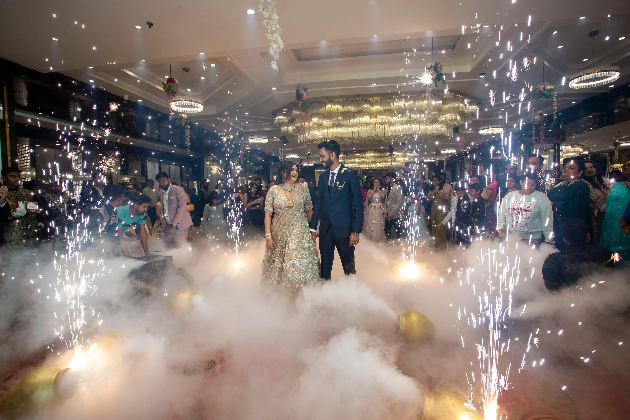 A couple dressed in traditional wedding attire dancing together on a decorated stage with fireworks and smoke, surrounded by guests at a celebration.