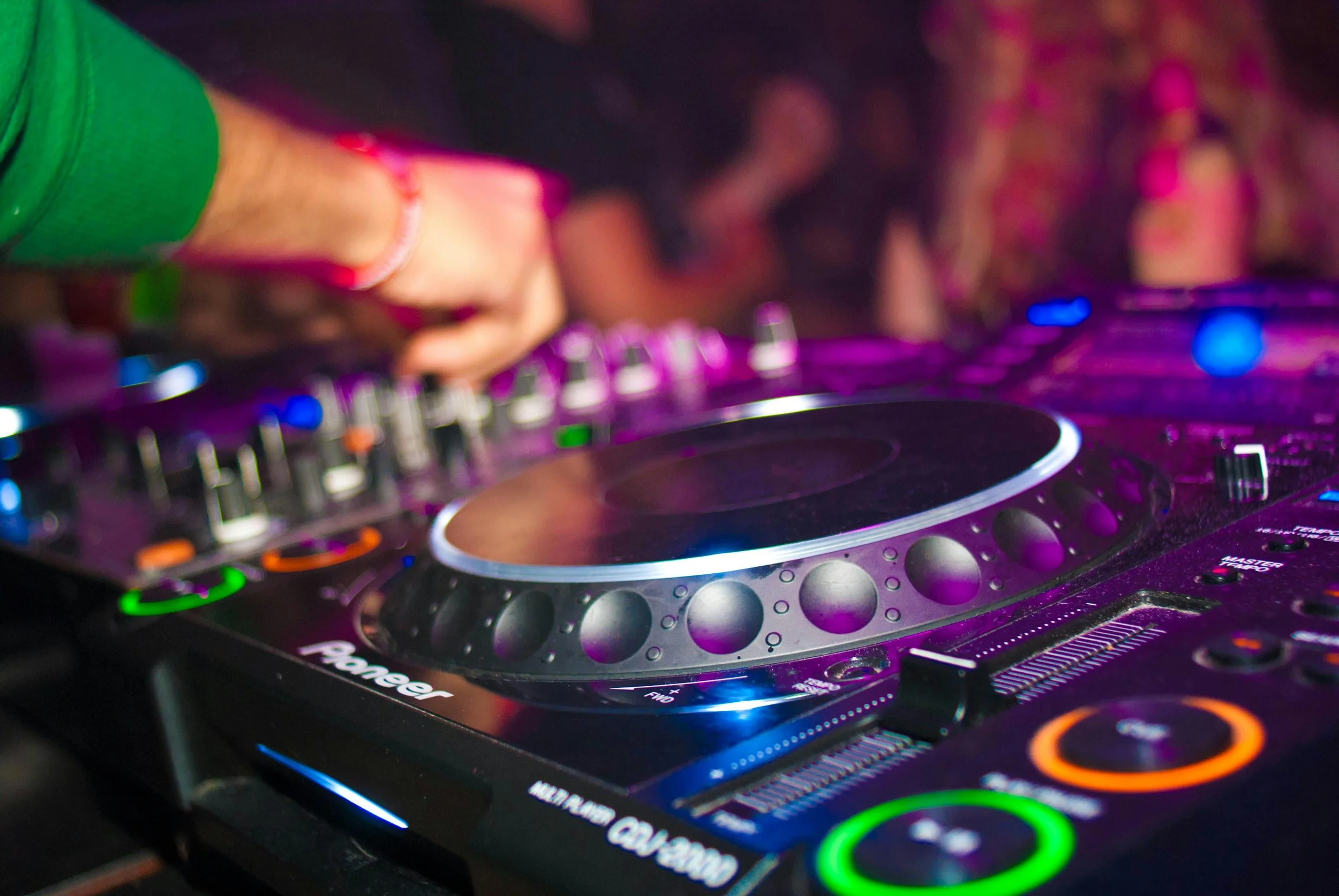A DJ's hand adjusting controls on a professional DJ mixer in a nightclub, with colorful lighting and a crowd in the background.