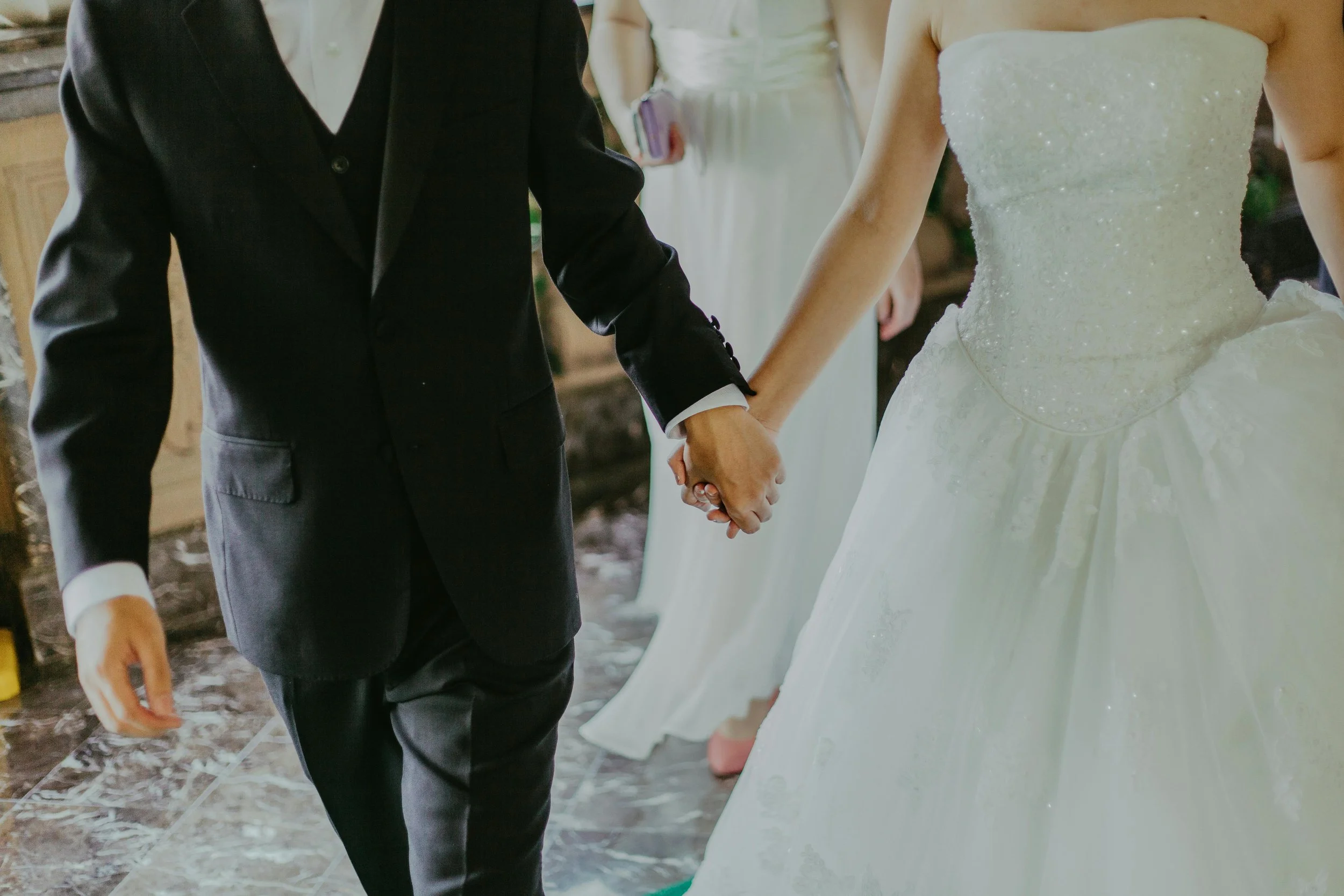 A couple holding hands during a wedding ceremony, with the bride in a white wedding dress and the groom in a black suit.