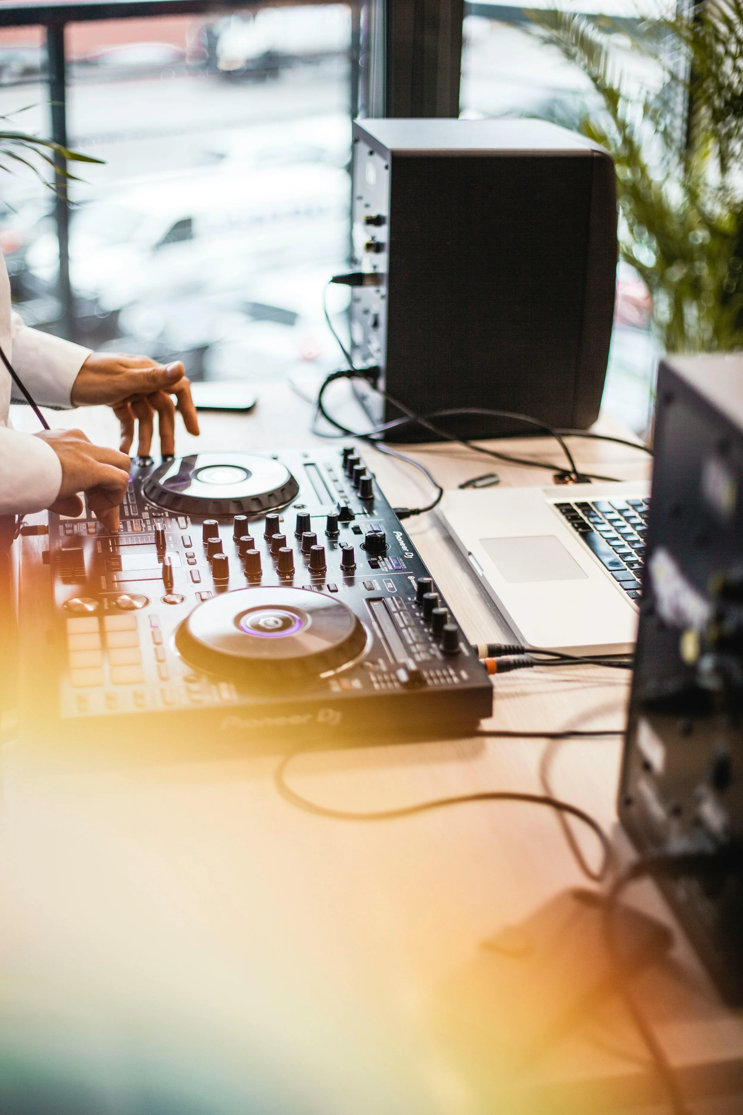 DJ mixing music at a club or event, with equipment including turntables, a laptop, and speakers on a wooden table near a window.