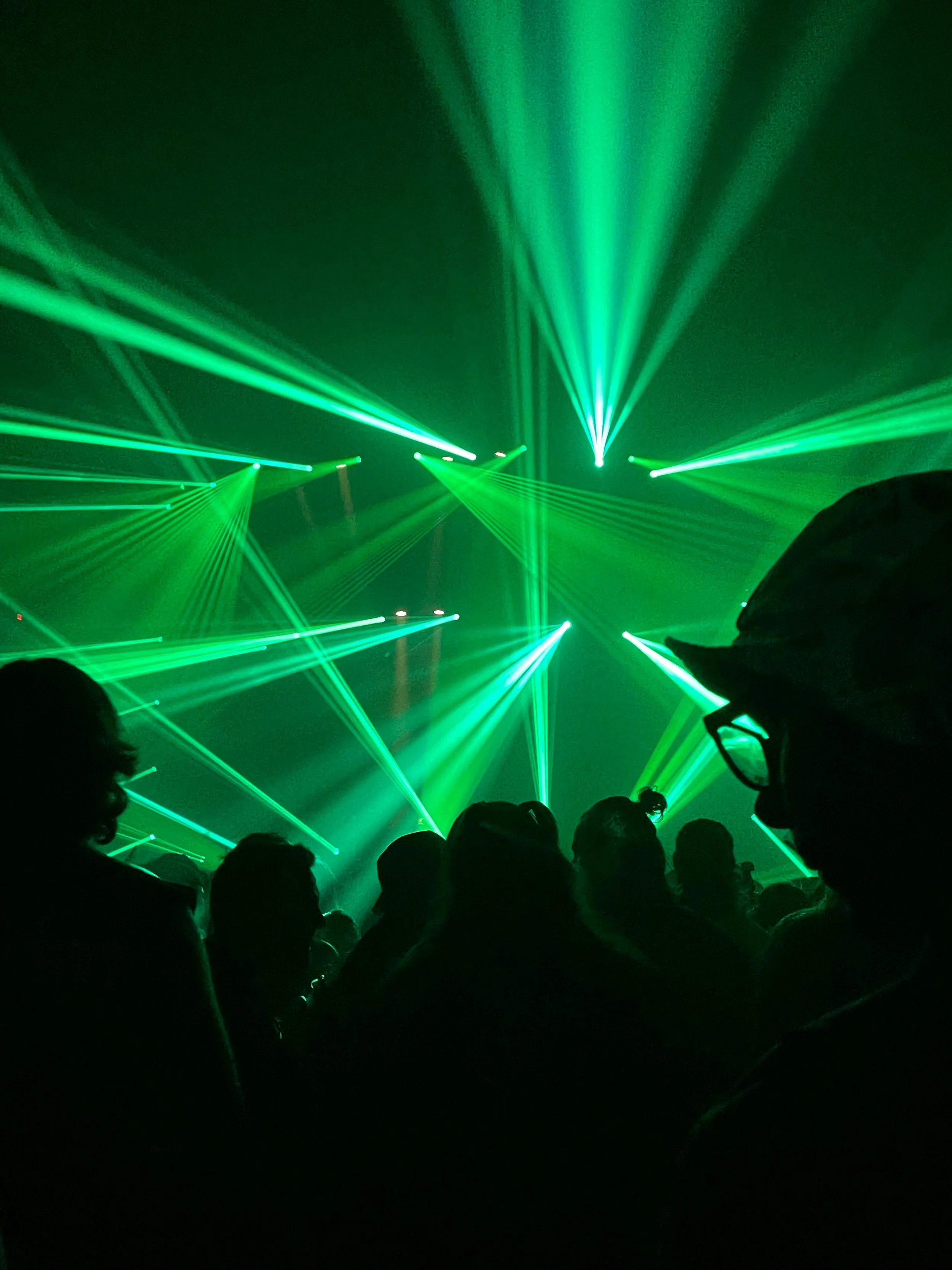 Dark concert scene with green laser lights creating patterns in the air, silhouettes of crowd members and some wearing glasses or glasses and hats.