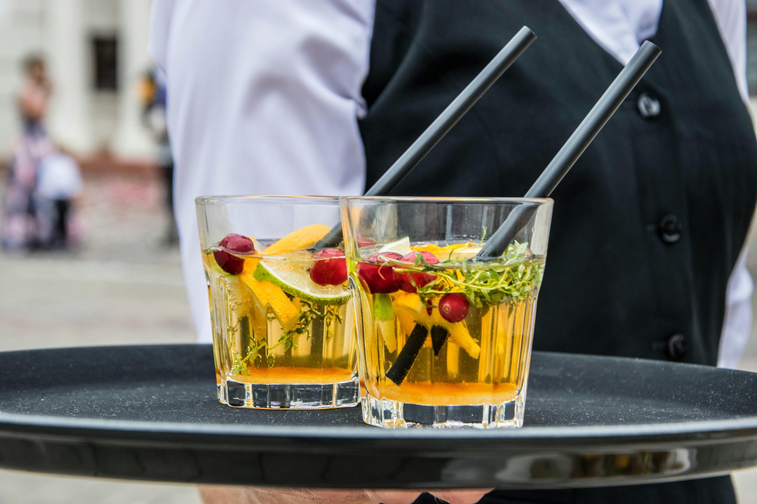 Two glasses of colorful fruit-infused water with lemon, lime, berries, and herbs on a black tray, held by a person in a black vest and white shirt.