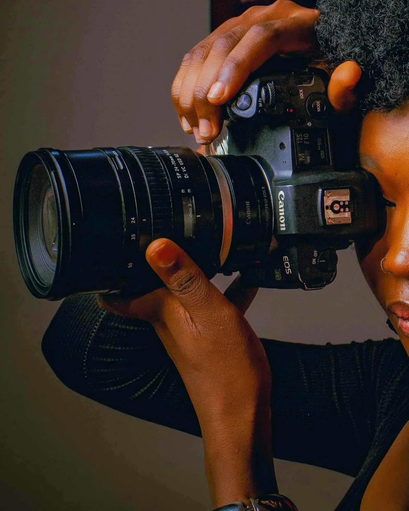 A person with dark skin and curly hair holding a Canon camera with a large lens, aiming it as if taking a photograph.