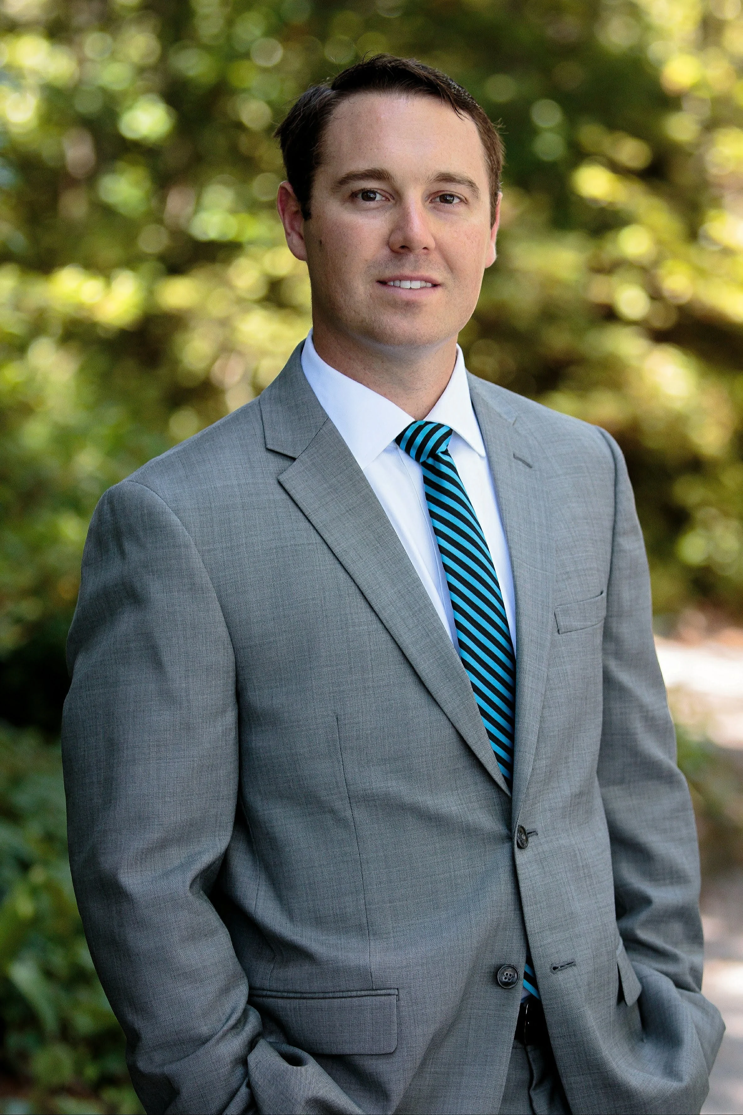 A man in a gray suit with a white shirt and blue striped tie standing outdoors in front of a blurred green background.