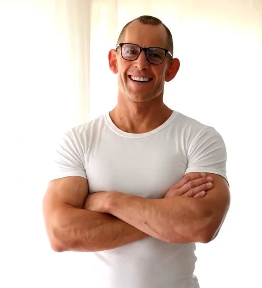 A smiling man with glasses, wearing a white T-shirt, standing with arms crossed in front of a light-colored background.