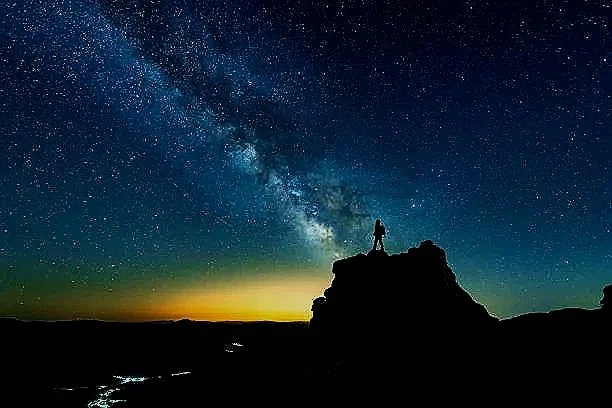 A person standing on a rock formation under a starry night sky with the Milky Way galaxy visible.