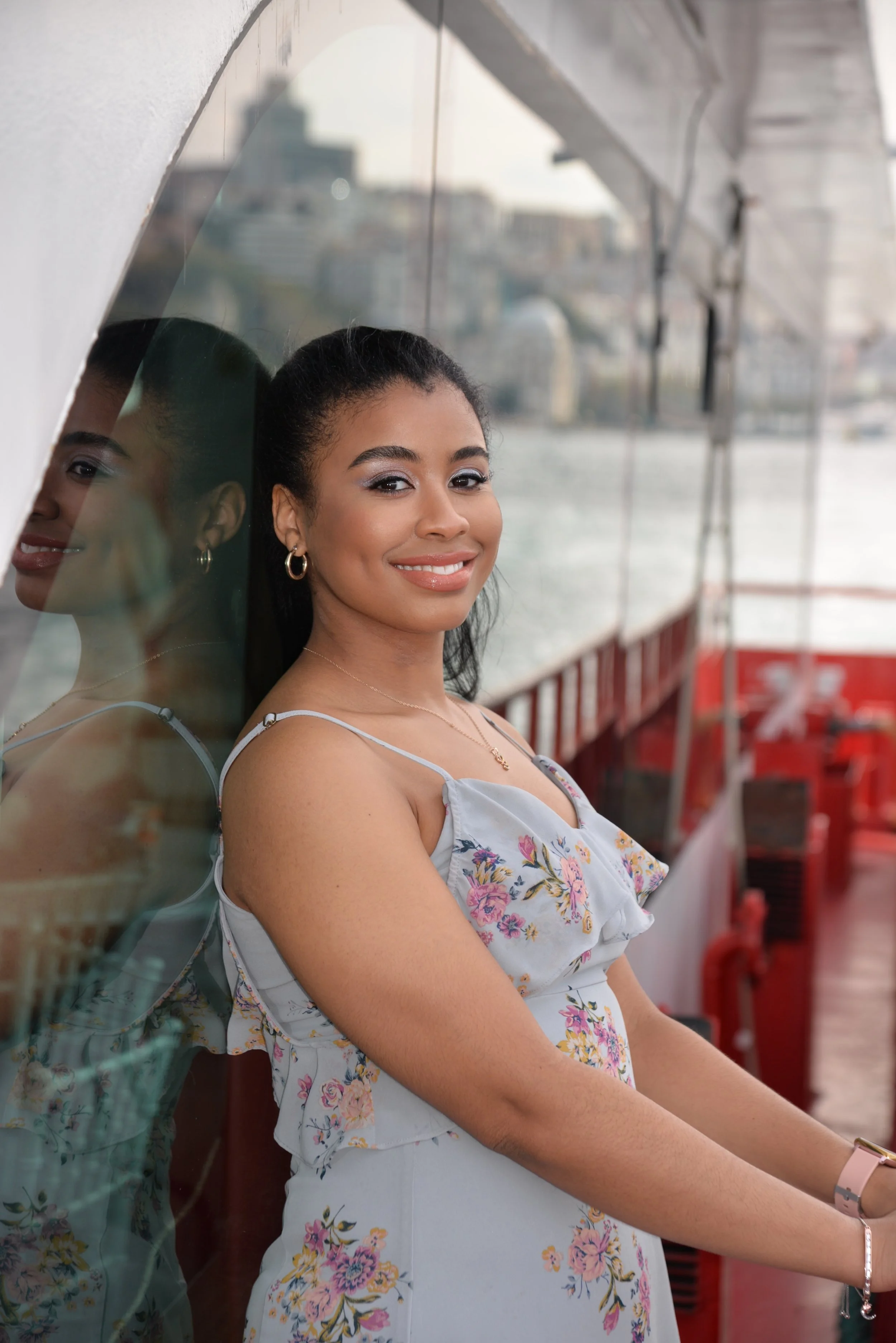 A woman with dark hair pulled back, wearing a white floral dress, leaning against a reflective surface, smiling, with a waterway and cityscape in the background.