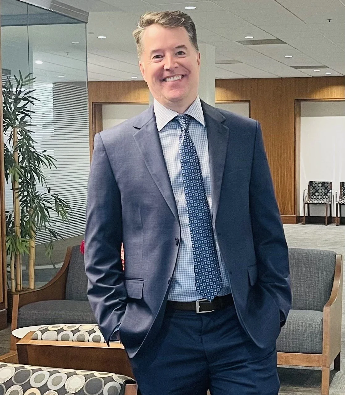 A smiling man in a business suit standing in an office lobby with wooden paneling and seating areas.