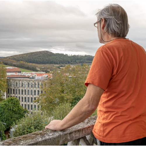 Een oudere man met een snor en bril zit op een houten bankje, naast een raam met een blauwe luik. Hij draagt een groene polo en kijkt vriendelijk naar de camera.