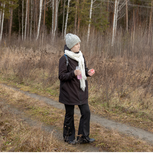 Portret van een oudere vrouw met witte haar, brillen en oorbellen, gekleed in een gele jurk met witte stippen, voor een witte tegelmuur.