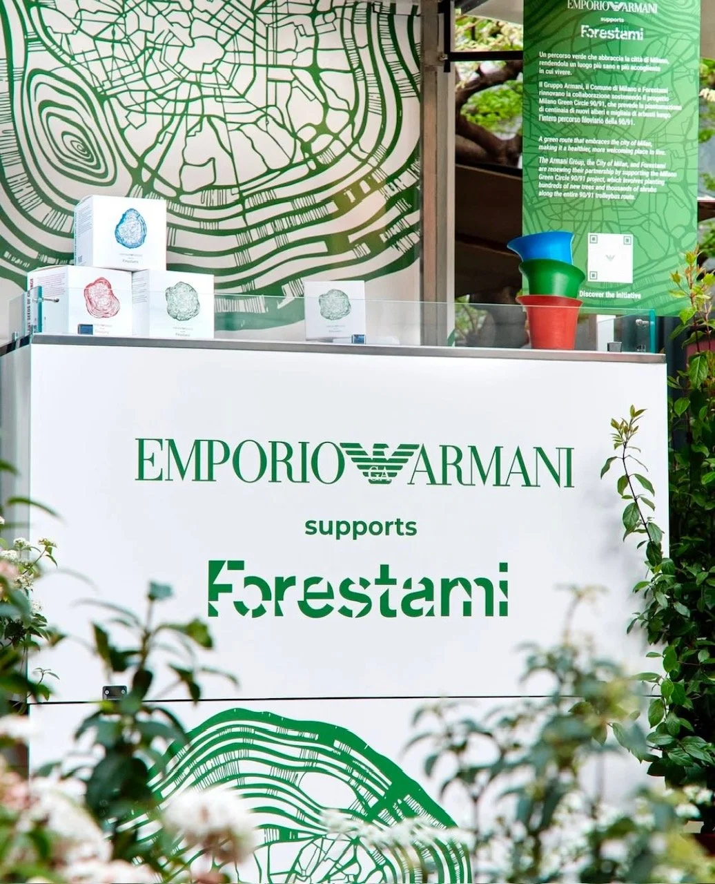 An event booth for Emporio Armani supporting Forestami, featuring a large white display with green text, informational signage, and stacked colored plant pots. The booth is decorated with green foliage.
