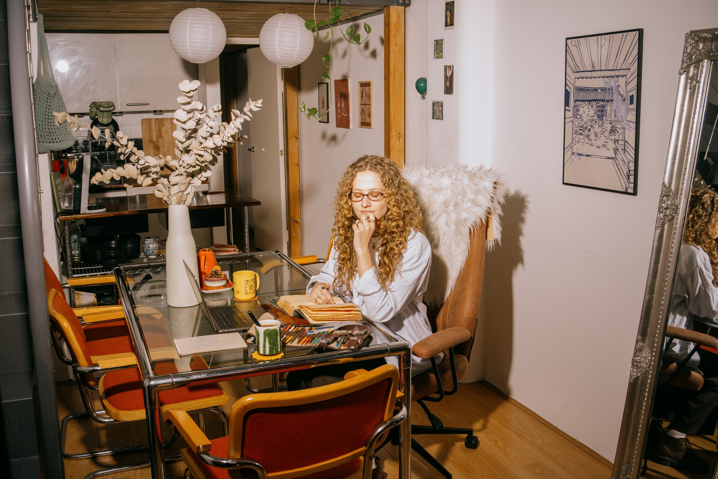 A woman with curly blonde hair and glasses sits at a cluttered desk in a cozy room, looking thoughtful with her chin resting on her hand. The desk has a laptop, notebooks, colored markers, and mugs. Behind her, a white wall with framed pictures and artwork. A large mirror reflects part of the room. The room has warm lighting and decorative paper lanterns hanging from the ceiling.