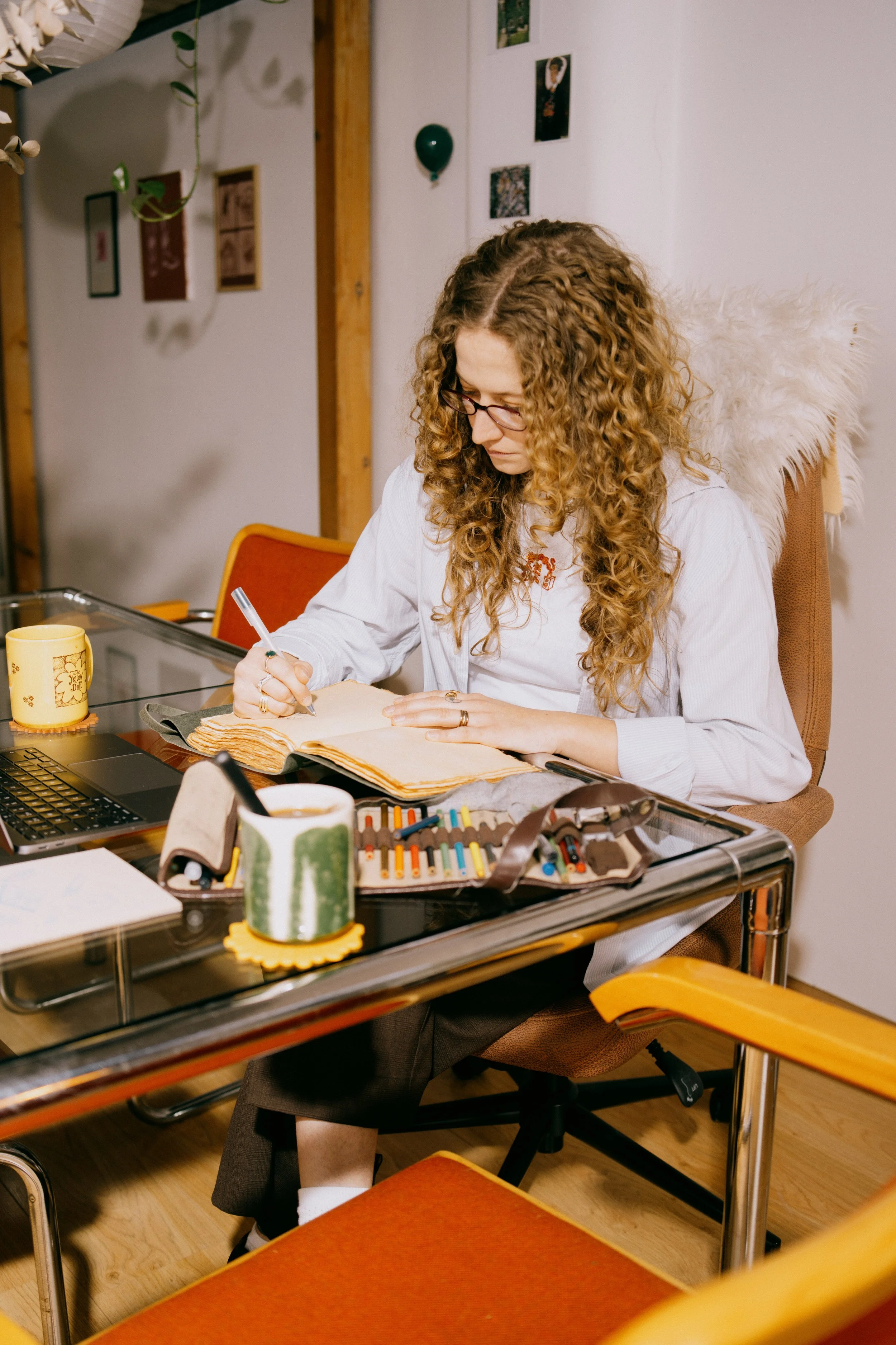 A woman with curly red hair and glasses sitting at a desk, writing in a large book with art supplies, a laptop, and coffee mugs around her. The room has wall decorations and a cozy, artistic atmosphere.
