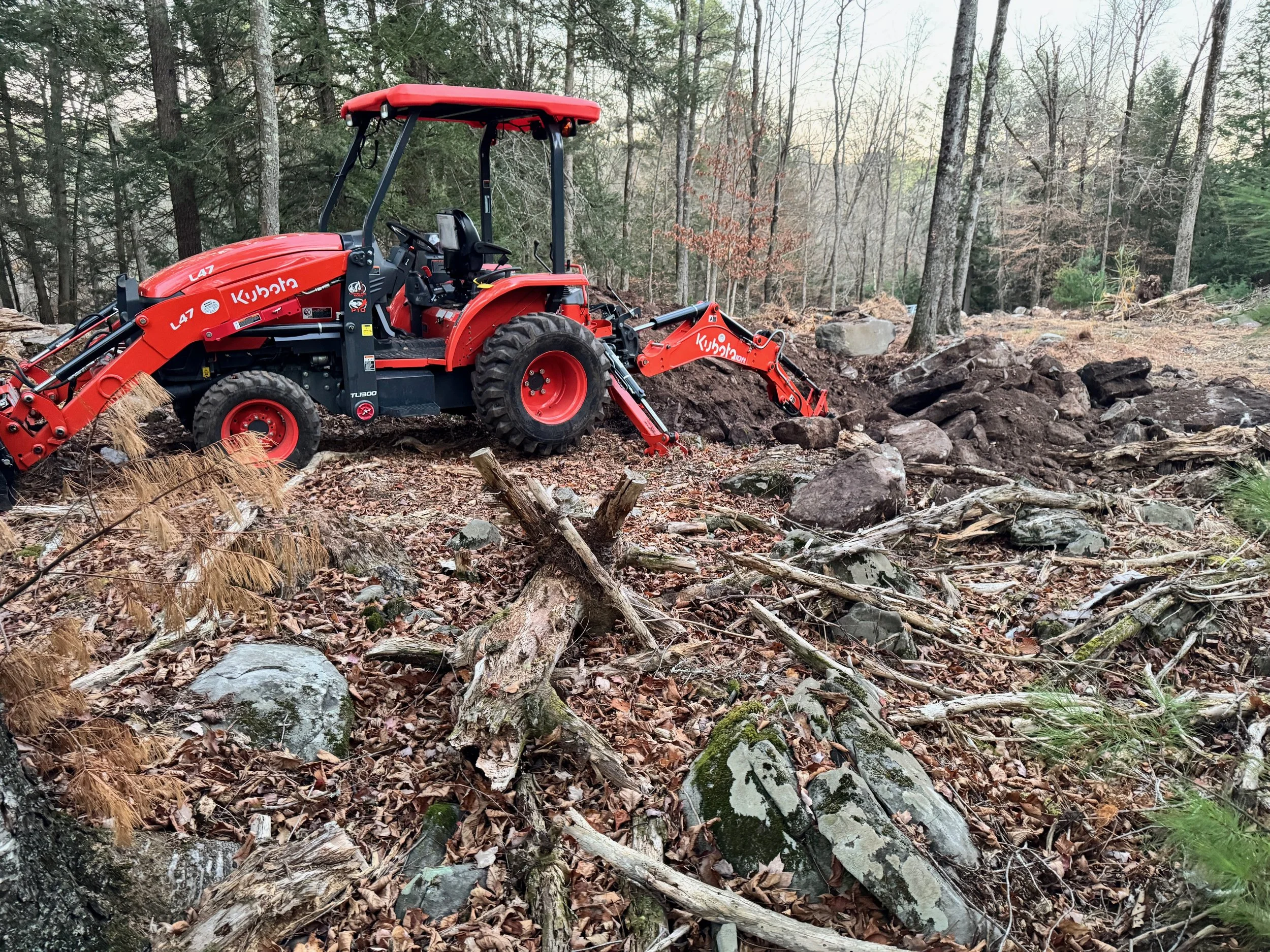 A Kubota L47 TLB  tractor digging a pond through large bouldery soils in a wooded area with rocks, fallen branches, and trees.