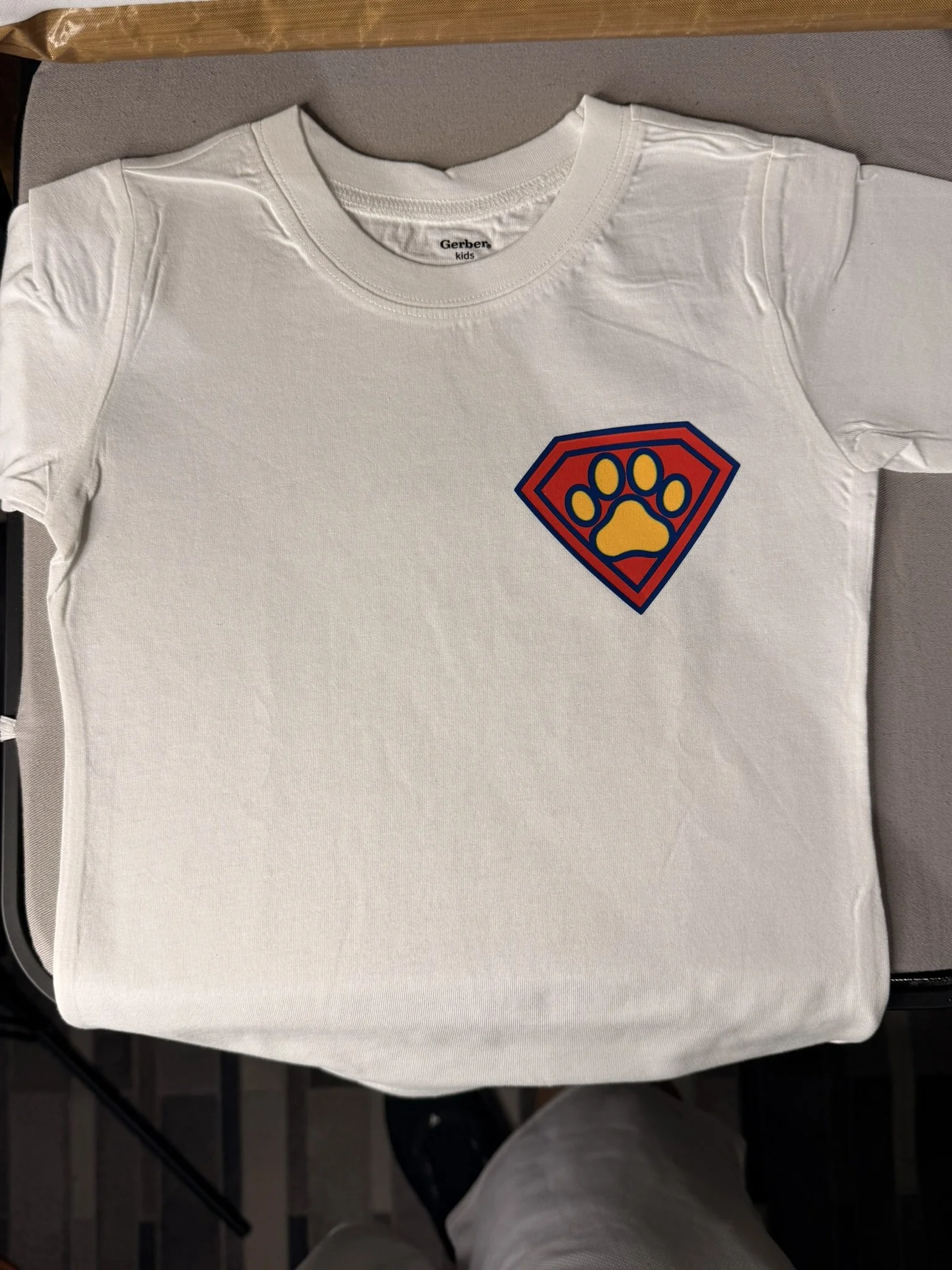 A white T-shirt with a superman-style emblem on the chest featuring a yellow paw print inside a blue and red shield outline.