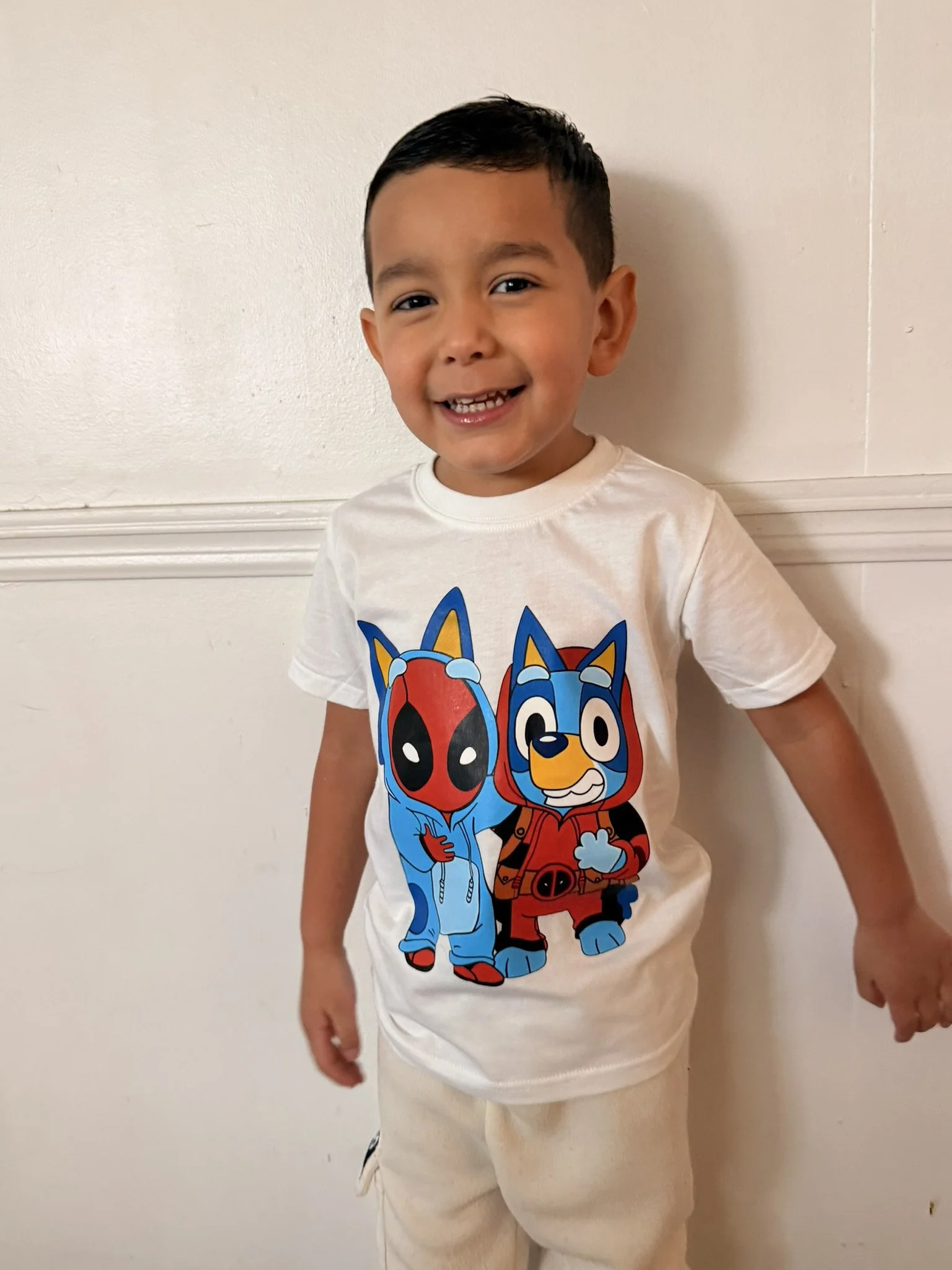 A young boy smiling and standing against a white wall, wearing a white T-shirt with cartoon characters Deadpool and a dog dressed as Deadpool.