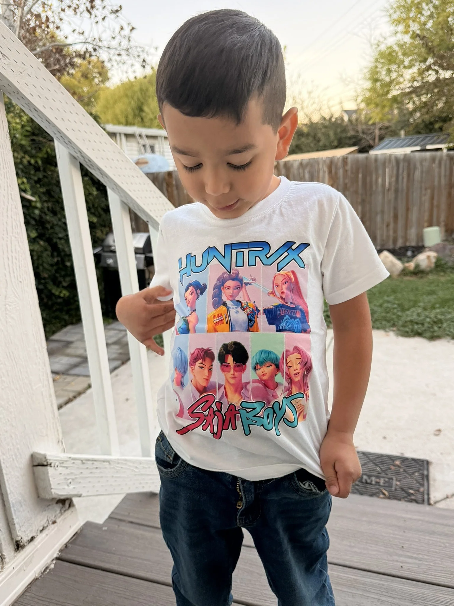Young boy wearing a white t-shirt with animated characters from the game 'HuntX' and 'Saia Boys' standing on a porch with a wooden fence, trees, and a backyard in the background.
