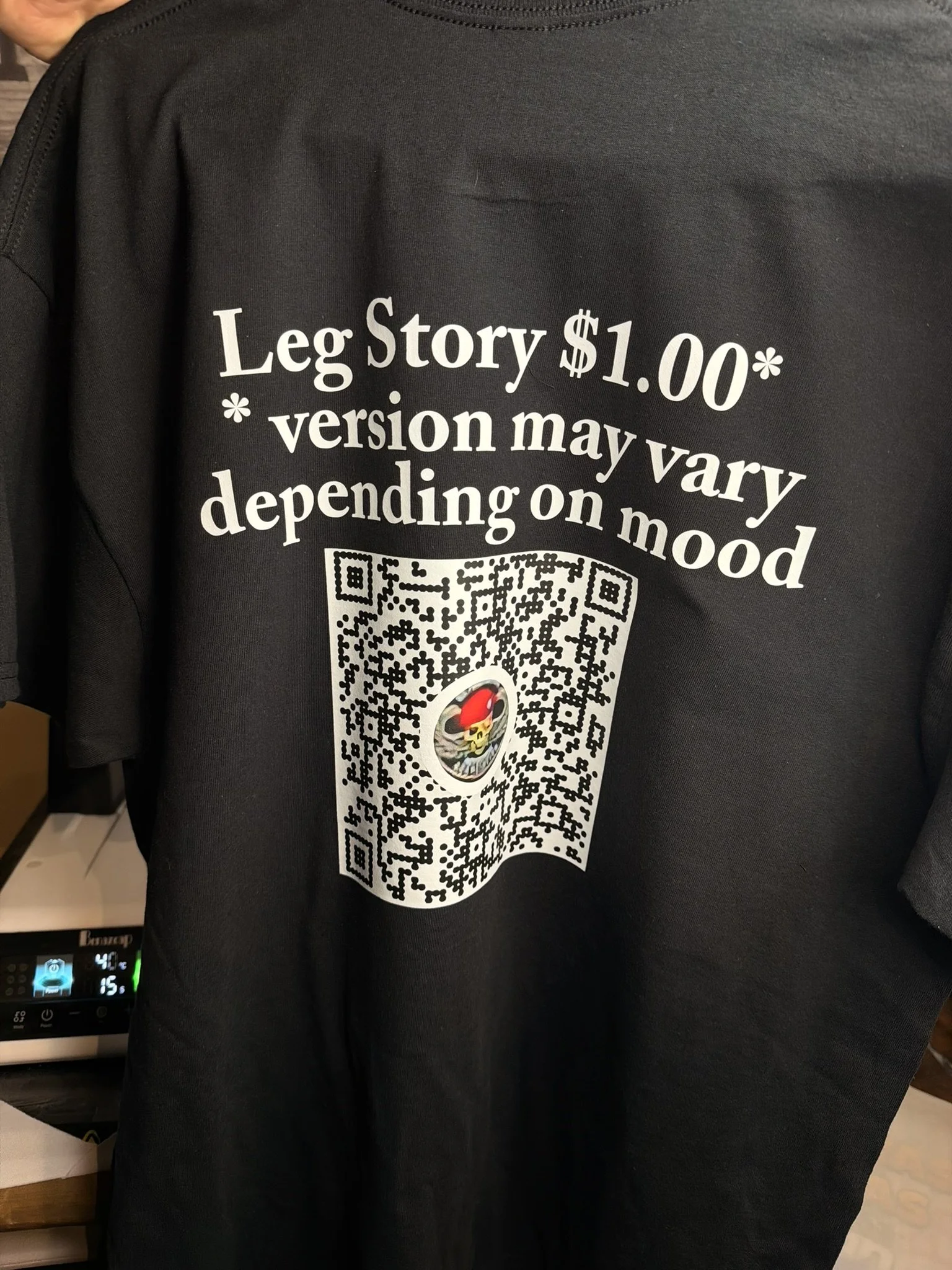 Black t-shirt advertising a Leg Story for $1.00, with a QR code and a logo in the center, hanging on a display rack.