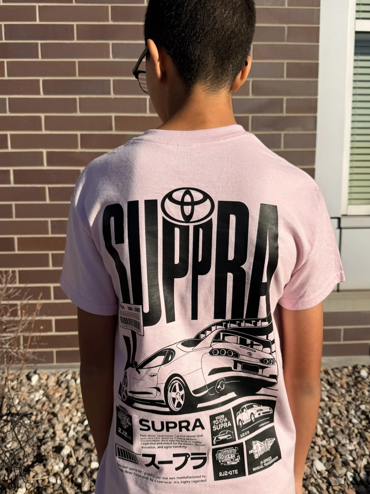 Back of a young person wearing a light pink T-shirt with black graphics of cars, the Toyota logo, and Japanese characters, standing outdoors with a brick wall and gravel ground.