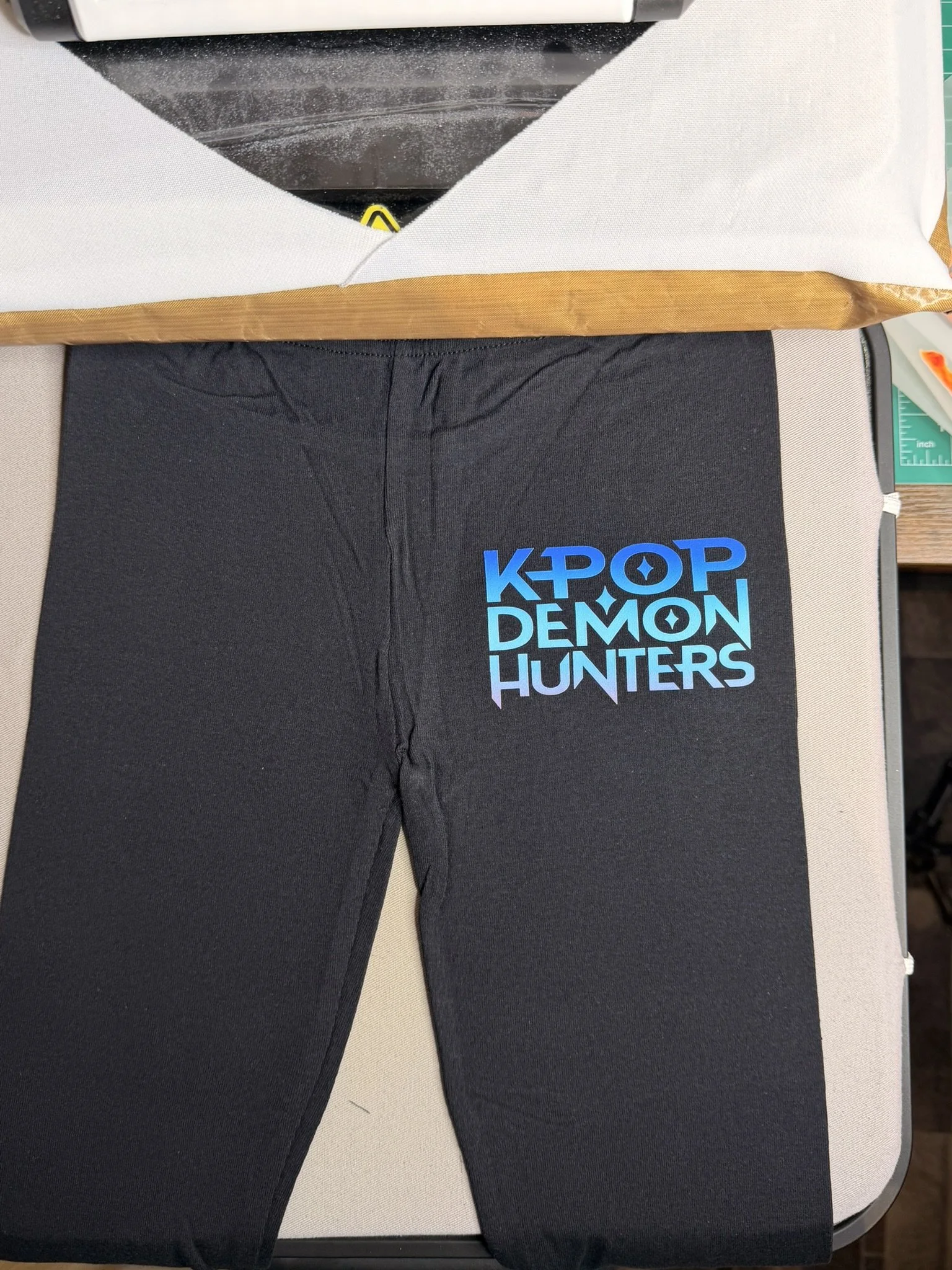 A pair of black leggings with a colorful 'KPOP DEMON HUNTERS' logo on the upper thigh. The leggings are placed on a light-colored cushioned surface, with a wooden item and other objects partially visible in the background.