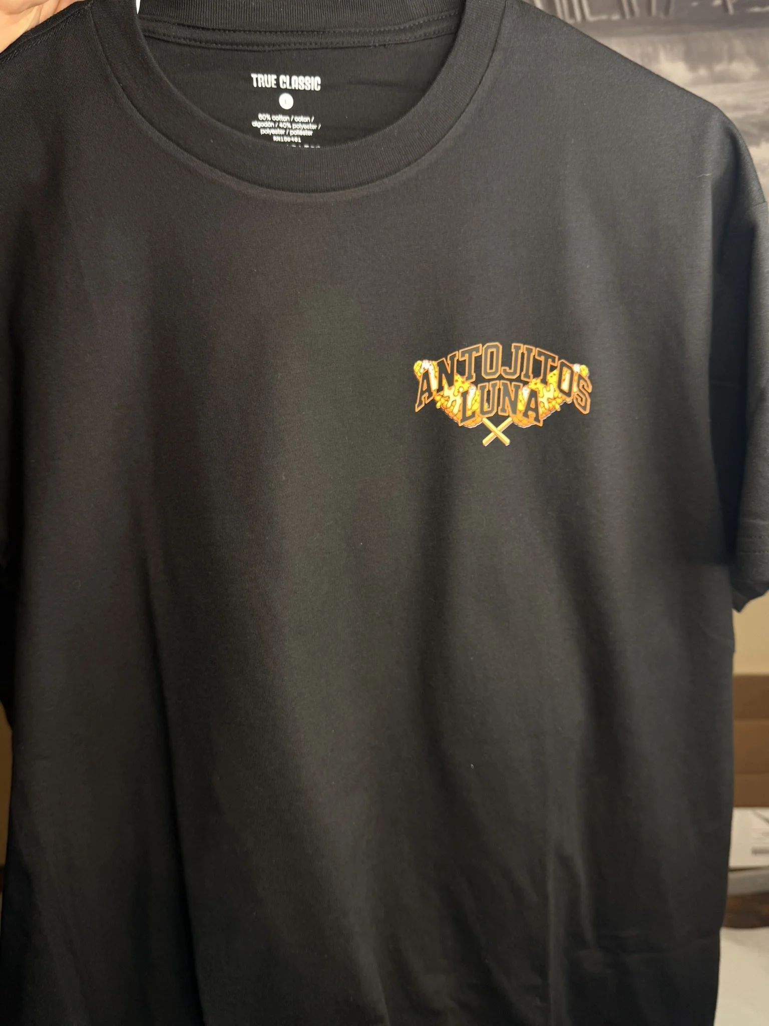 Black T-shirt with a small printed logo on the left chest area that reads "Antojitos Luna" in yellow and orange lettering with a decorative design.