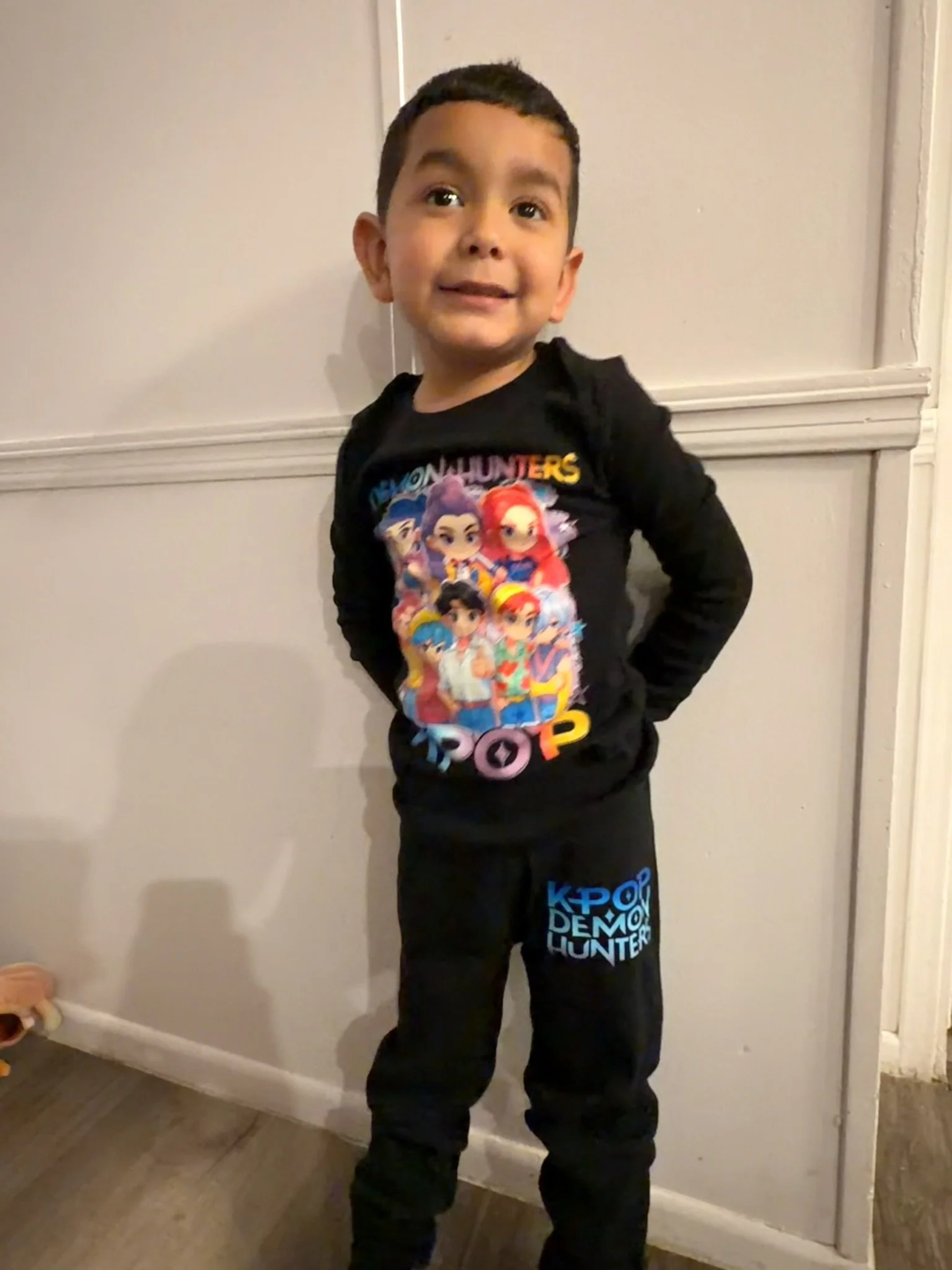 A young boy with short dark hair standing indoors against a light-colored wall. He is wearing a black "Dream Hunters" long-sleeved shirt with colorful characters and matching black pants with a Blue "K-pop Demon Hunter" logo. He has a cheerful expres