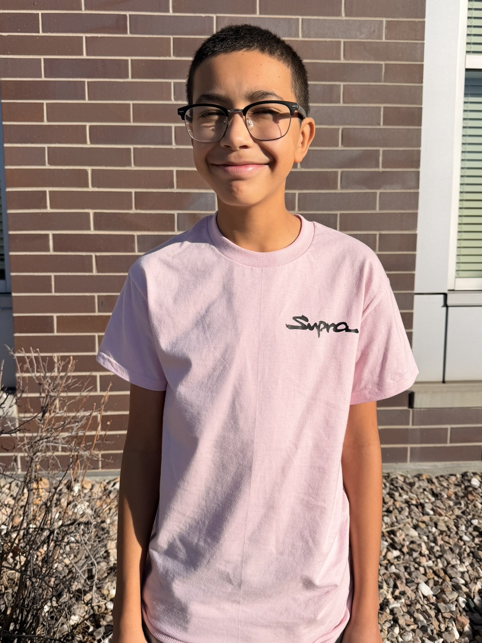 A smiling boy wearing glasses and a pink Supreme t-shirt stands outside in the sunlight, in front of a brick wall and a window.