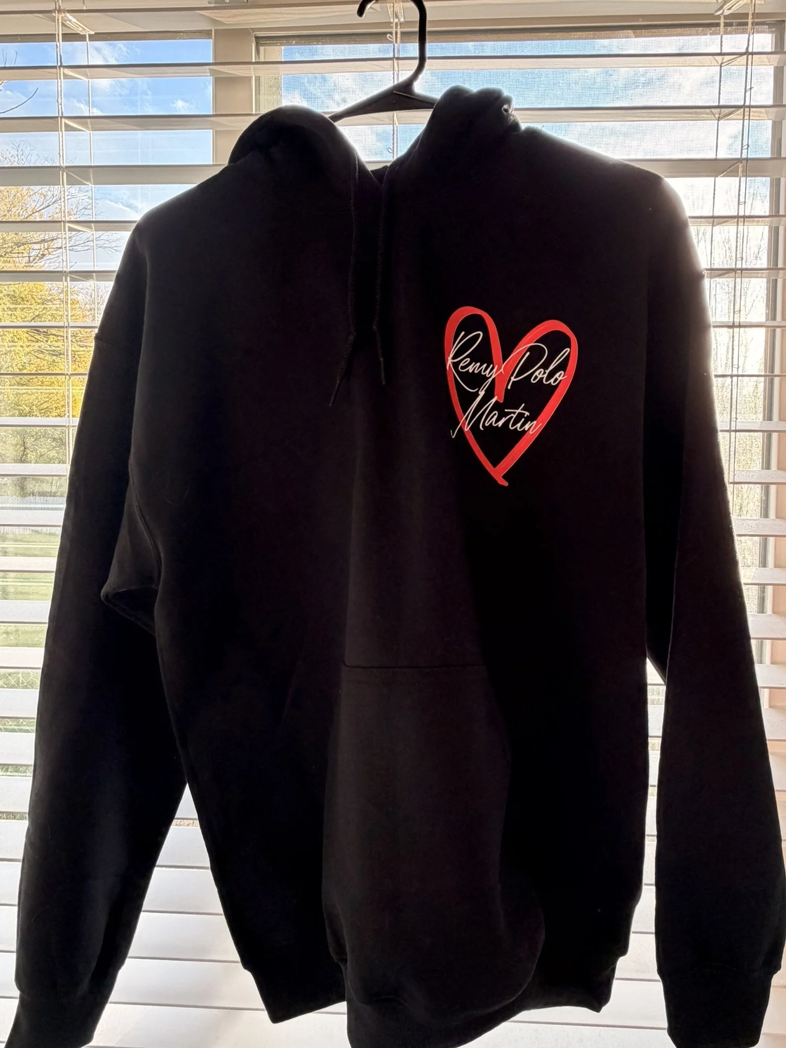 Black hoodie with a red heart design and the text "Remy Polo Martin" written inside the heart, hanging on a hanger in front of window blinds.