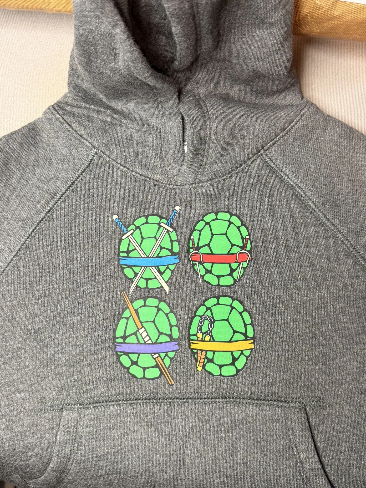 Gray hoodie with four green turtle shells designed with different ninja weapons, including swords, nunchucks, and a bo staff.