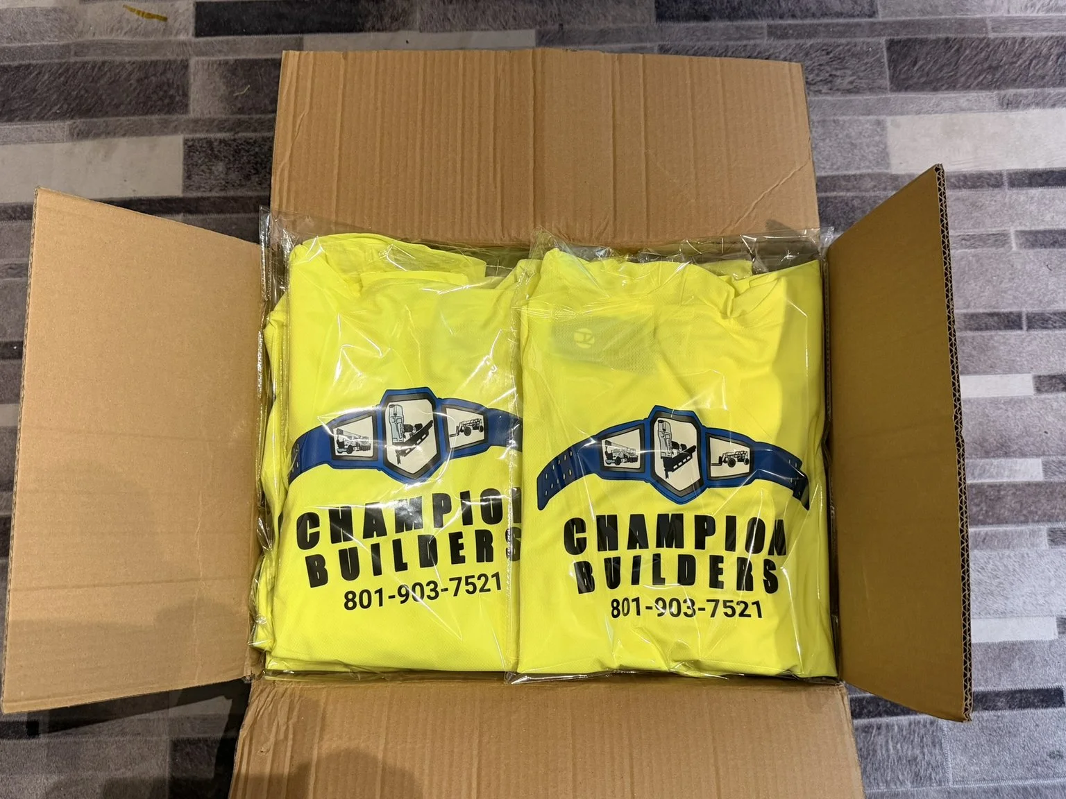 Open cardboard box containing yellow bags with 'Champion Builders' logo and phone number, on gray tile floor.
