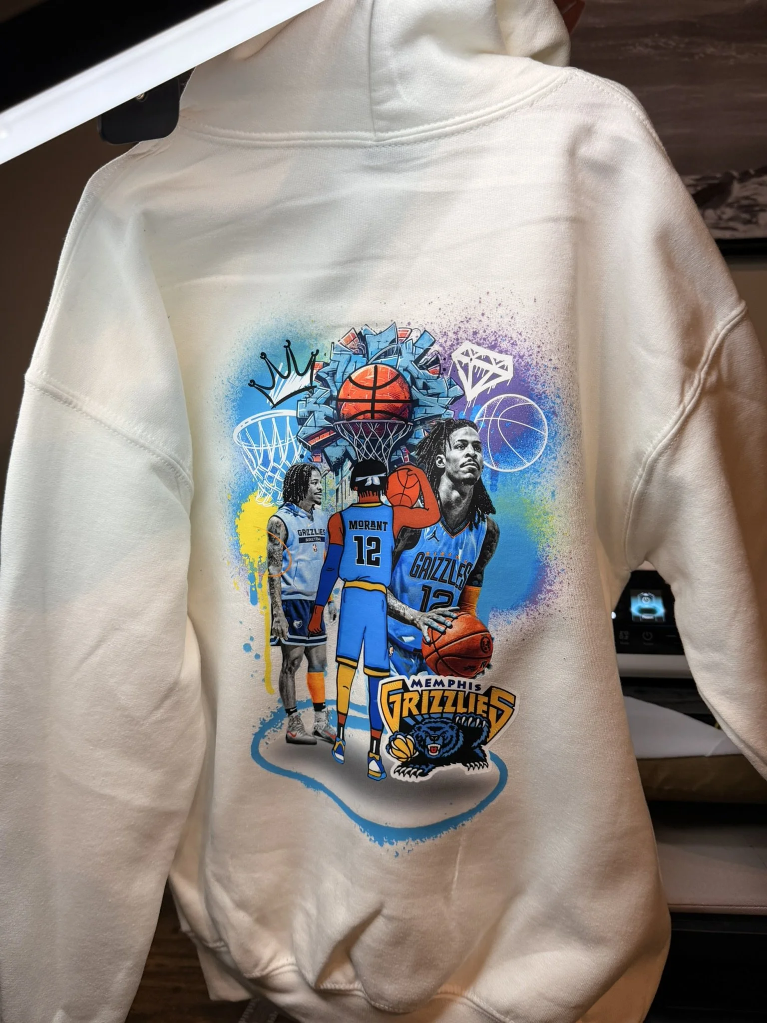 A white hoodie featuring a vibrant graphic of a Memphis Grizzlies basketball player, multiple images of the same player, the team logo, and various basketball-themed illustrations.