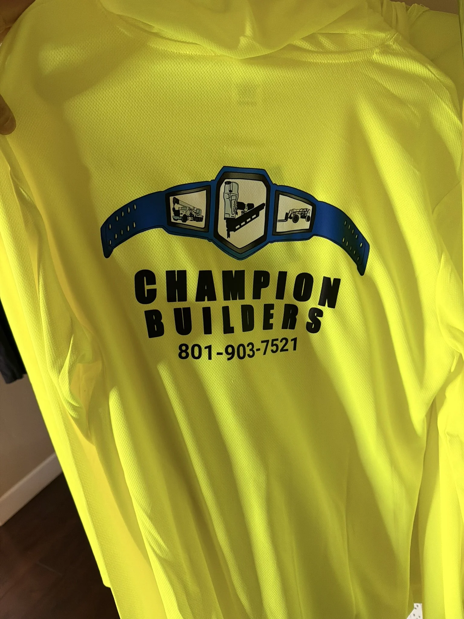 Bright yellow work vest with a logo for 'Champion Builders' including icons of a crane, a construction worker, and a forklift, along with the phone number 801-903-7521.