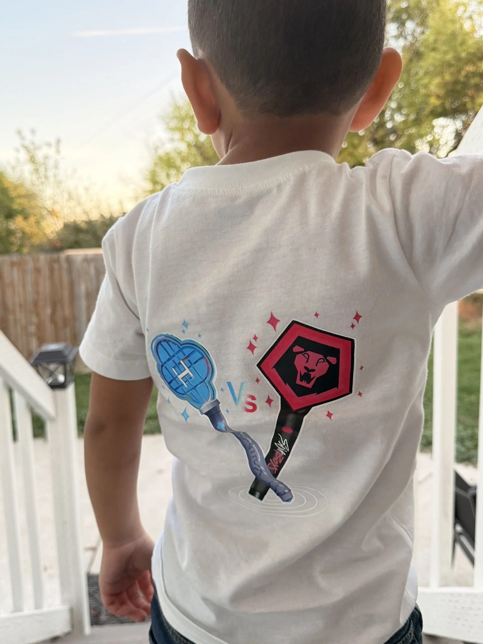 Back of a young boy wearing a white T-shirt with a graphic design of two paddles and the text 'VS', standing outdoors on a porch with a grassy yard and trees in the background.