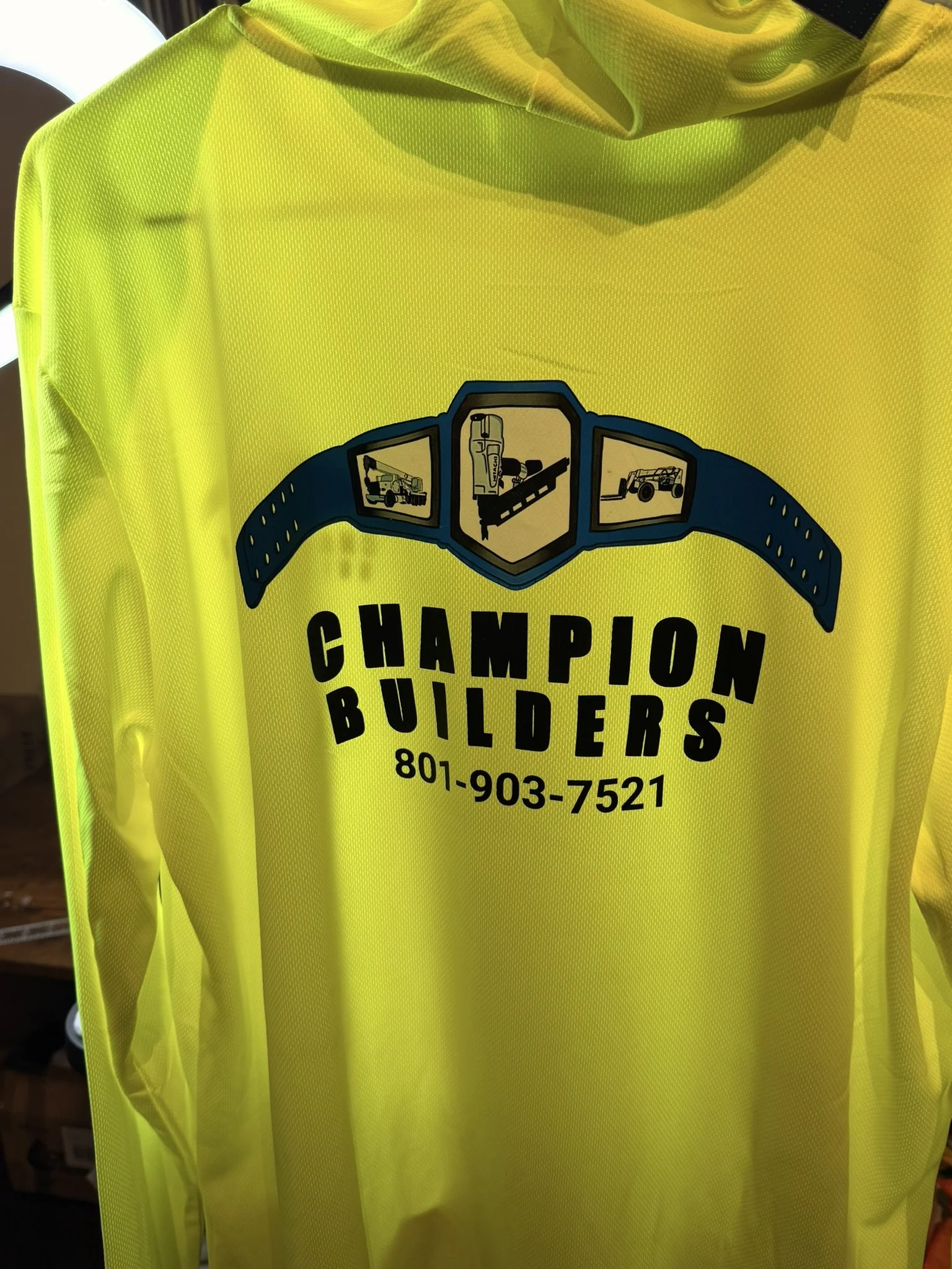 Bright yellow construction safety vest with a logo for Champion Builders, featuring images of construction equipment and tools, and a phone number 801-903-7521.
