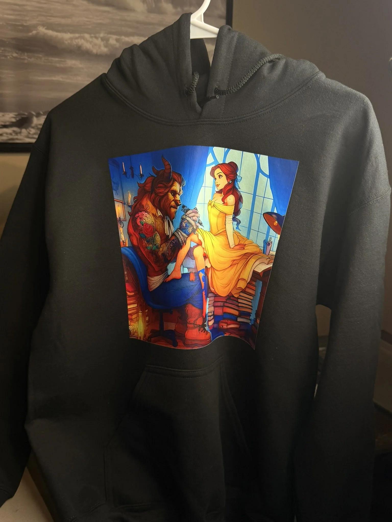 A black hoodie featuring a colorful illustration of the Beast from Beauty and the Beast tattooing a young woman with red hair in a yellow dress, inside a room with a large window.